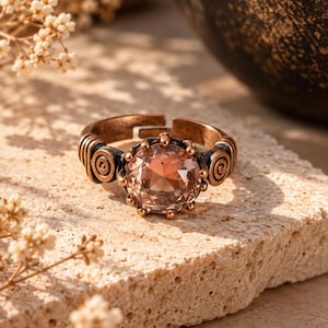 May include: An adjustable copper-toned ring featuring a large, round, faceted peach-colored gemstone set in a prong setting. The band is decorated with embossed spiral patterns on both sides of the central stone and has an open back design.
