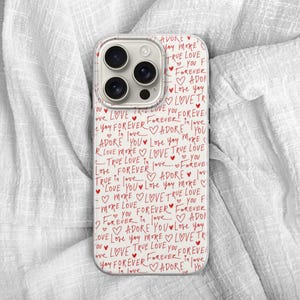May include: A white phone case with a repeating pattern of red handwritten text and hearts. The words "Love," "Adore," and "Forever" are visible. The phone case is on a white, textured surface.