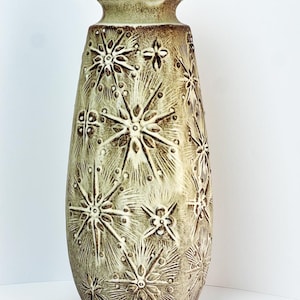Puede incluir: Un jarrón alto de cerámica verde oliva con superficie texturizada y base acampanada. Presenta un patrón repetitivo de estrellas estilizadas y diseños florales en marrón oscuro. El borde tiene un patrón geométrico.