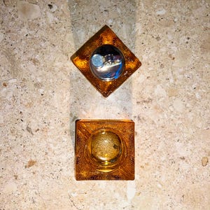 Pode incluir: Duas cinzeiros de vidro âmbar. Um é quadrado com uma reentrância circular, o outro em forma de diamante, segurando um isqueiro prateado. Ambos estão em uma superfície salpicada marrom-claro.