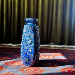 Puede incluir: Un jarrón azul cobalto con una superficie texturizada y un patrón de estrella. El jarrón tiene una base ancha y un cuello estrecho. El jarrón está sobre una alfombra estampada con diseños rojos y azules.