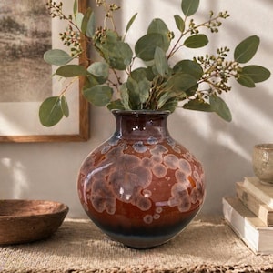 May include: A round ceramic vase with a glossy finish featuring a deep reddish-brown base color and lighter, crystalline-like circular patterns in shades of tan and grey. The vase has a narrow neck and holds a sprig of green eucalyptus branches.