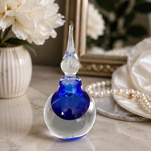 May include: A clear glass perfume bottle with a spherical base and a deep blue glass insert. The bottle features a tall, slender, clear glass stopper that tapers to a point, creating a distinct contrast between the transparent and blue glass elements.