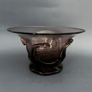 May include: A vintage amethyst-colored glass bowl. The bowl has a wide, shallow shape with a flared rim and decorative handles. The base is a hexagonal shape. The glass has a glossy finish.