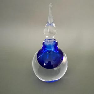 Può includere: Un flacone di profumo in vetro trasparente con base blu e tappo trasparente allungato. Il flacone ha una forma rotonda e bulbosa con una fascia blu al centro. Il tappo è lungo e appuntito.