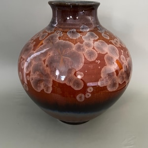 May include: A round ceramic vase with a glossy finish, featuring a deep reddish-brown base colour that transitions to a darker tone at the bottom. The surface is decorated with light-coloured, circular crystalline patterns scattered across the body.