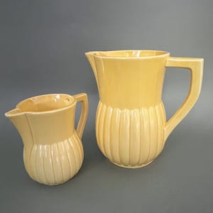 Peut inclure: Deux pichets en céramique jaune. Le plus grand pichet a une anse haute et rectangulaire et des détails verticaux côtelés sur le corps. Le plus petit pichet a un design similaire. Les deux ont un bec verseur.
