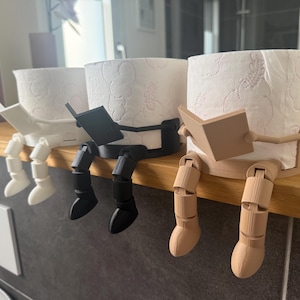 Könnte beinhalten: Drei Toilettenpapierhalter in Weiß, Schwarz und Beige. Jeder Halter zeigt eine Figur mit beweglichen Beinen, die über die Kante hängen, und Armen, die ein kleines Buch halten. Die Halter sind so konzipiert, dass sie um eine Rolle Toilettenpapier passen.