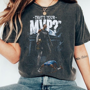 Pode incluir: Camiseta cinza escuro com um jogador de basquete segurando um troféu e o texto "THAT'S YOUR MVP?" em branco. O gráfico inclui um jogador caído e um edifício ao fundo. A camiseta é de estilo vintage.
