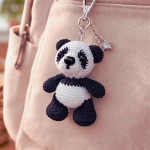 May include: A handmade crochet panda bear keychain. The panda has a black and white color scheme, with black accents on the ears, eyes, and limbs. It is attached to a silver-colored clasp with a small star charm.