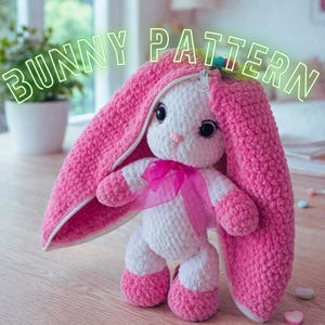 May include: A handmade crochet bunny with a pink zippered hood and matching pink accents on the feet and hands. The bunny has a white body, black eyes, and a pink ribbon. The text "BUNNY PATTERN" is displayed above the bunny.