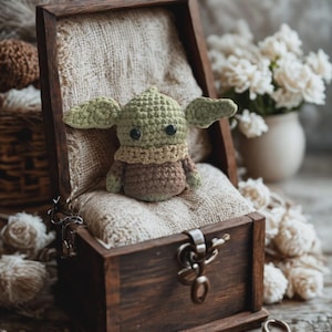 Op de afbeelding: Een handgemaakte gehaakte figuur van een personage met grote oren, zittend in een kleine, donkere houten schatkist. De figuur is groen en bruin, met zwarte ogen. De kist is gevoerd met beige stof en heeft een metalen sluiting.