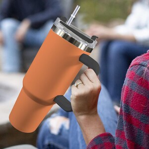 May include: An orange insulated tumbler with a black handle and a clear lid with a straw. The tumbler is held by a person wearing a plaid shirt. The tumbler is designed to keep drinks cold or hot.