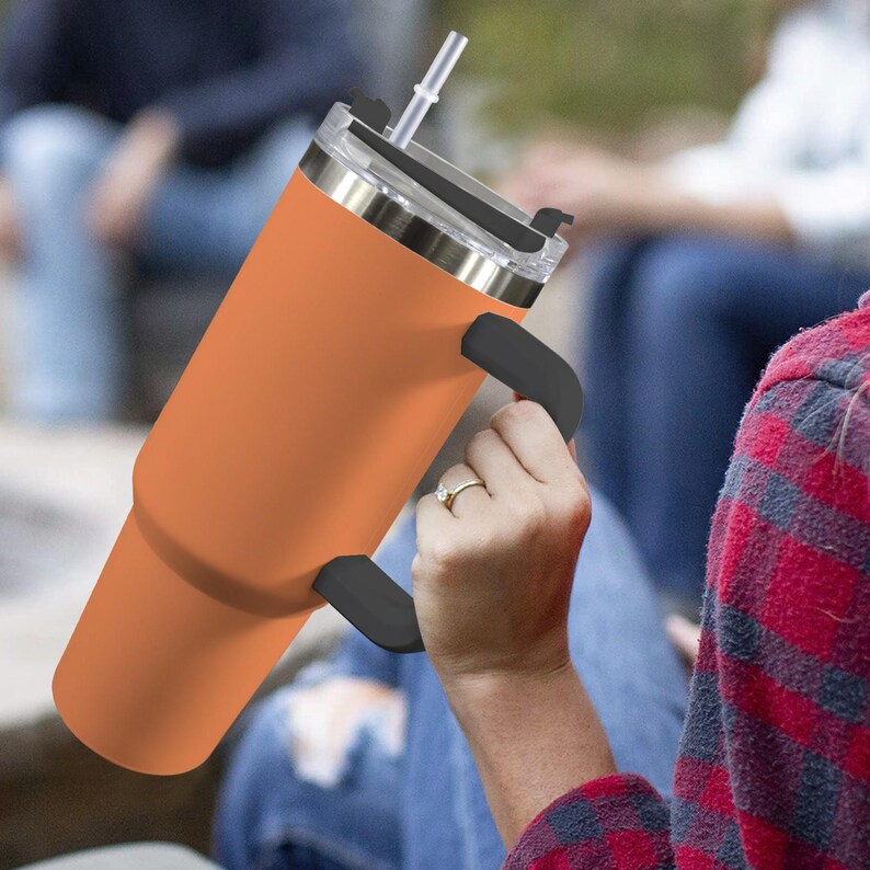 May include: An orange insulated tumbler with a black handle and a clear lid with a straw. The tumbler is held by a person wearing a plaid shirt. The tumbler is designed to keep drinks cold or hot.