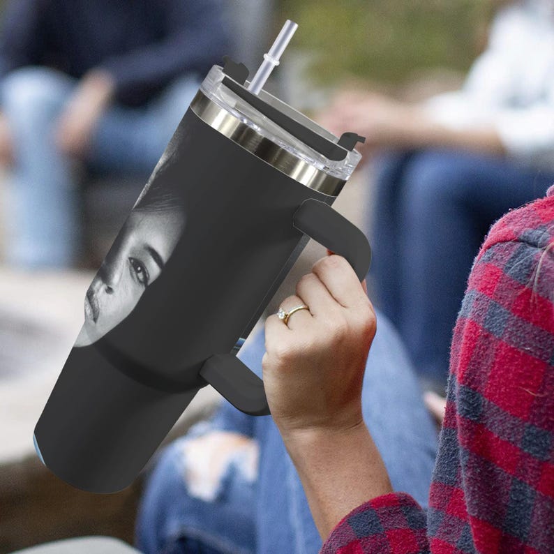 May include: Black insulated tumbler with a silver rim, black handle, and a clear lid with a straw. The tumbler features a black and white image of a person's face. The tumbler is held by a person wearing a ring.