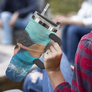 May include: A stainless steel tumbler with a black handle and a clear lid, featuring a vibrant image of a person paddling a canoe in blue water. The tumbler includes a straw for easy drinking.