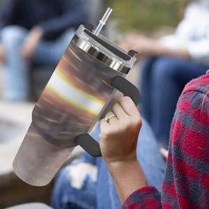 K&ouml;nnte beinhalten: Ein Edelstahl-Reisebecher mit schwarzem Griff und Deckel sowie einem durchsichtigen Strohhalm. Der Becher hat ein wirbelndes Design in Braun-, Gelb- und Wei&szlig;t&ouml;nen. Die Person, die den Becher h&auml;lt, tr&auml;gt ein rot-schwarz kariertes Hemd.