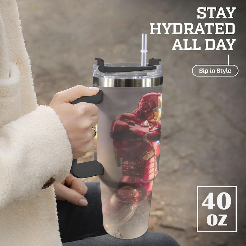K&ouml;nnte beinhalten: Edelstahlbecher mit schwarzem Griff und Deckel, mit einer Iron Man-Grafik. Der Becher hat einen durchsichtigen Strohhalm und den Text "Stay Hydrated All Day" und "Sip in Style". Er fasst etwa 1,18 Liter.