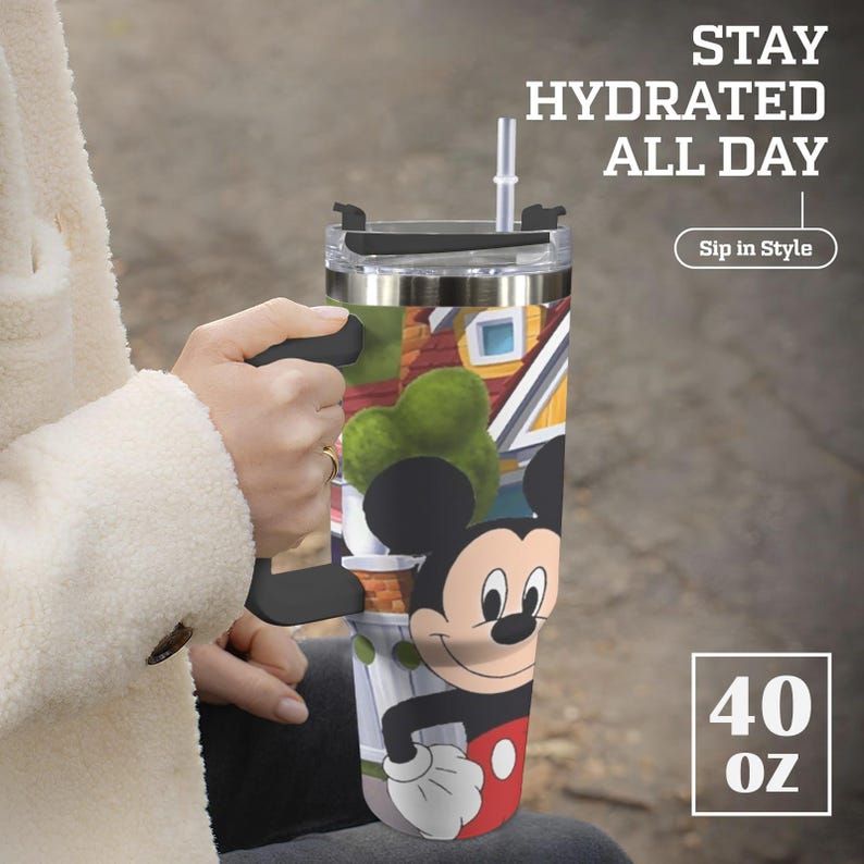 May include: A 40 oz insulated tumbler featuring a Mickey Mouse design. The tumbler has a black handle, a clear lid, and a straw. The design includes a colorful illustration of a town scene. Text on the tumbler reads "Stay Hydrated All Day" and "Sip in Style."