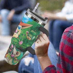 May include: A stainless steel tumbler with a black handle and straw. The tumbler features a colorful graphic of cartoon characters in a forest setting with a blue sky. The tumbler is held by a person.