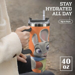 May include: A stainless steel tumbler with a cartoon image of a gray bunny with purple eyes and a carrot. The tumbler is orange and has a black handle and lid. Text on the tumbler reads "Stay Hydrated All Day" and "Sip in Style". The tumbler holds 40 oz.