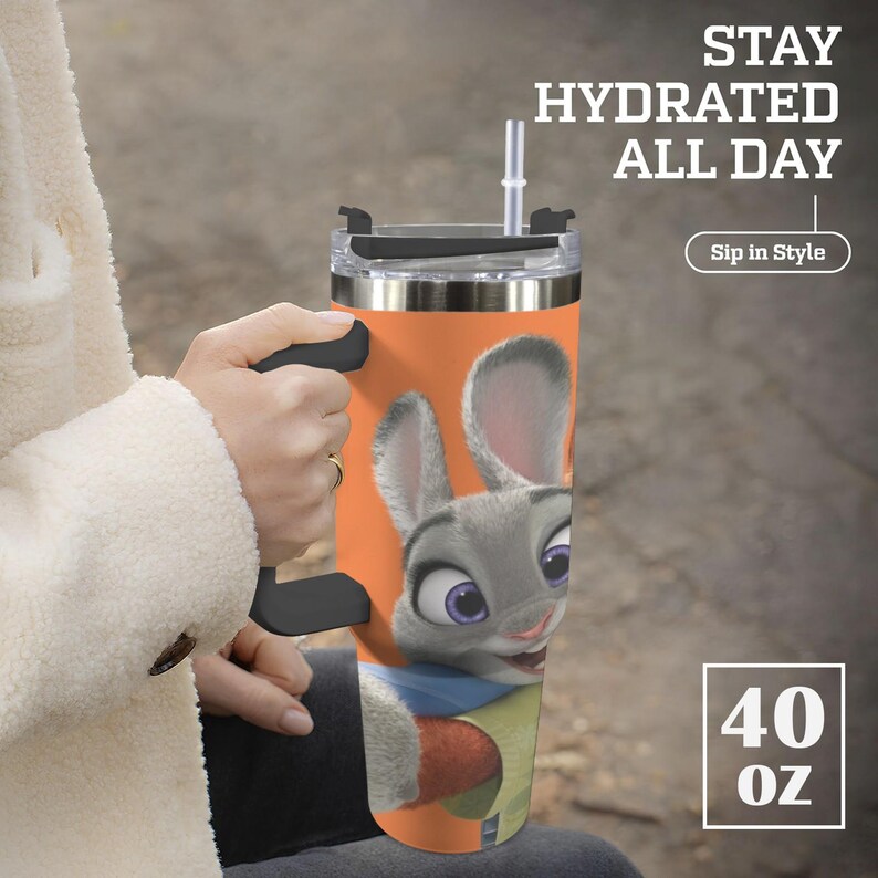May include: A stainless steel tumbler with a cartoon image of a gray bunny with purple eyes and a carrot. The tumbler is orange and has a black handle and lid. Text on the tumbler reads "Stay Hydrated All Day" and "Sip in Style". The tumbler holds 40 oz.