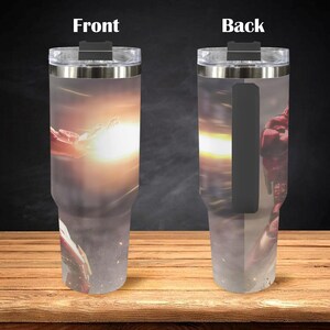 K&ouml;nnte beinhalten: Ein Edelstahlbecher mit Superhelden-Motiv. Die Vorderseite des Bechers zeigt eine dynamische Szene mit hellem Licht, w&auml;hrend die R&uuml;ckseite eine Figur in einem roten Anzug zeigt. Der Becher hat einen schwarzen Griff und einen transparenten Deckel. H&ouml;he: ca. 24 cm.