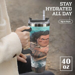 May include: A 40 oz tumbler featuring a cartoon image of Maui from the movie Moana. The tumbler has a black handle and lid, with a clear straw. Text on the tumbler reads "Stay Hydrated All Day" and "Sip in Style".
