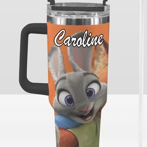 May include: A stainless steel travel mug with a black handle and lid. The mug features a cartoon image of Judy Hopps from Zootopia, with the name "Caroline" in white script. The background is orange, and a clear straw is included.