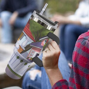 May include: A stainless steel travel tumbler with a black handle and a clear lid with a straw. The tumbler features a colorful cartoon illustration of houses, trees, and a white picket fence.