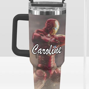 K&ouml;nnte beinhalten: Ein Edelstahl-Reisebecher mit einer rot-goldenen Iron Man-Grafik. Der Becher hat einen schwarzen Griff und Deckel, und der Name "Caroline" ist in wei&szlig;er Schrift auf der Vorderseite geschrieben. Ein klarer Strohhalm ist enthalten.
