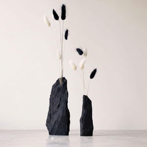 May include: Two black vases with a rough, rock-like texture. The taller vase on the left holds three black and two white dried bunny tail grasses. The shorter vase on the right holds two white and one black dried bunny tail grass stem.