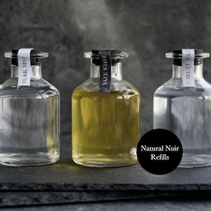 May include: Three clear glass bottles with black stoppers and white labels. The labels read "PEAK MIST", "NAZE STONE", and "FOREST SAGE". The center bottle contains a yellow liquid. A black circle with the text "Natural Noir Refills" is in the foreground.