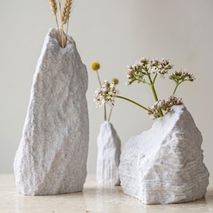 May include: Three white stone-textured vases with irregular, jagged shapes. The tallest vase holds dried wheat stalks, the middle vase holds a single yellow flower, and the shortest, widest vase holds a cluster of small white flowers with green stems.