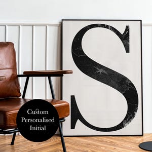 Puede incluir: Una impresión en blanco y negro enmarcada con una gran letra "S" con un diseño texturizado. La obra de arte se muestra junto a un sillón de cuero marrón con patas de metal negro. La imagen también incluye una lámpara de pie negra y el texto "Custom Personalised Initial".