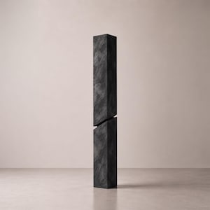 Puede incluir: Una escultura alta y rectangular de color gris oscuro con una textura rugosa similar a la piedra. El objeto está compuesto por dos segmentos verticales separados por una brecha diagonal en el medio, creando una apariencia dividida.