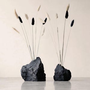 May include: Two black, rock-like textured vases of different sizes, each holding a collection of thin, tall stems topped with fluffy white and black seed heads. The vases have a rough, uneven surface and irregular, jagged shapes.