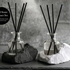 May include: A Dual Stone Diffuser Set featuring two clear glass bottles with black reed sticks. Each bottle rests in a natural stone base, one dark grey and one light grey. The text on the image reads: Dual Stone Diffuser Set, 2x forms. Includes bottles and reeds for each.