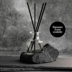 May include: A clear glass diffuser bottle with black reeds, set on a dark grey stone. The bottle rests on a black slate tray. A black circle with the text "Signature scents available" is visible.