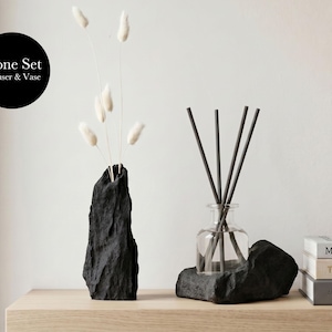May include: A black stone set, comprising a vase with dried white flowers and a diffuser with reeds. The vase and diffuser are on a wooden surface. The text "Stone Set Diffuser & Vase" is in a black circle.
