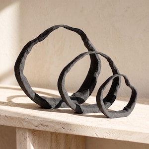 May include: Three black circular sculptures of varying sizes arranged in a row. Each sculpture features a thick, uneven, and textured surface with a matte finish, forming an open, organic ring shape that stands upright on a flat base.