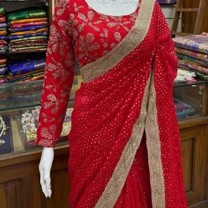 May include: A vibrant red saree with intricate gold embroidery and a matching blouse. The saree features a delicate pattern and a gold border. The long-sleeved blouse has detailed floral patterns. A mannequin displays the saree with a necklace.