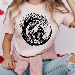May include: A light pink t-shirt featuring a black graphic design. The design includes a tree, the Earth, three silhouetted figures with raised fists, flowers, and stars. The shirt has short sleeves.