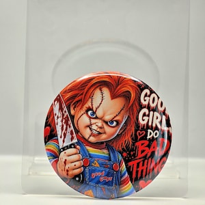 May include: A circular pinback button featuring a graphic of a red-haired doll with stitches on its face, holding a blood-stained knife. The doll wears blue denim overalls over a striped shirt. The text reads GOOD GIRLS DO BAD THINGS.