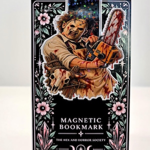 May include: A rectangular magnetic bookmark featuring a horror-themed illustration of a masked figure holding a blood-stained chainsaw. The black background is decorated with pink flowers, crystals, and the text "MAGNETIC BOOKMARK THE HEX AND HORROR SOCIETY".