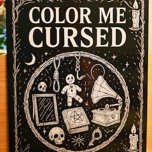 May include: A black book cover with the title "COLOR ME CURSED" in white lettering. The cover features a circular design with a noose, voodoo doll, skulls, and a gramophone. The author's name, AMANDA FARRELL, is at the bottom.