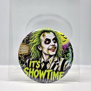 May include: A circular pinback button featuring a graphic illustration of a character with wild green hair, pale skin, and a black and white striped suit. The text BEETLEJUICE appears on a sign, and the words IT'S SHOWTIME are printed in green.