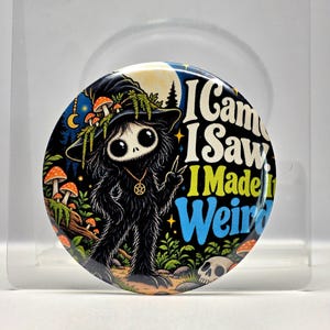 May include: A circular pinback button featuring a black, furry creature with large eyes wearing a witch hat adorned with mushrooms. The text reads I Came I Saw I Made It Weird. The background shows a forest scene with a moon, stars, and a skull.