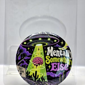 May include: A circular pinback button featuring a colorful illustration of a UFO beaming up a pink brain in a desert landscape with cacti and purple clouds. The text reads Mentally Somewhere Else and a sign says OUT OF HERE with an alien icon.