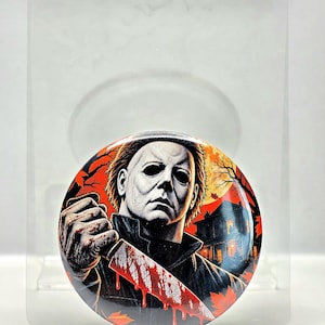 May include: A circular pinback button featuring a graphic illustration of a masked figure with dark hair, wearing a dark jacket, and holding a large knife covered in red blood. The background depicts a house at night under a large orange moon.